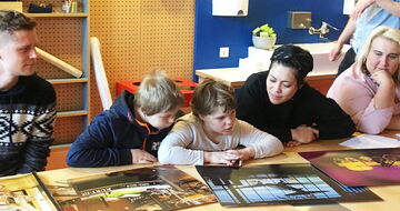 Zwei Schüler*innen betrachten zusammen mit einer Einzellhelferin große Fotos am Tisch
